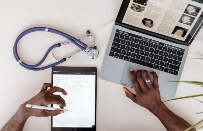 A doctor using a tablet and laptop with a stethescope on their desk