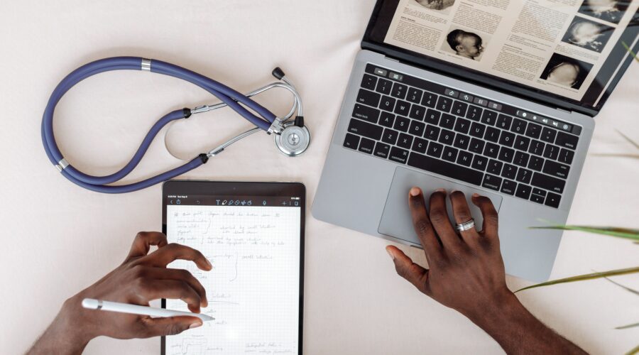 A doctor using a tablet and laptop with a stethescope on their desk