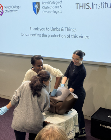 Three clinicians in gloves practising disimpaction on a childbirth training simulator, in front of a screen showing the Royal College of Midwives, Royal College of Obstetricians and Gynaecologists and THIS Institute logos.