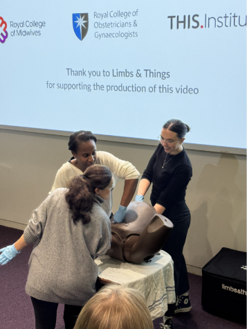 Three clinicians in gloves practising disimpaction on a childbirth training simulator, in front of a screen showing the Royal College of Midwives, Royal College of Obstetricians and Gynaecologists and THIS Institute logos.