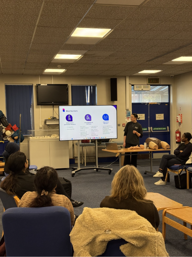 A trainer presents a slide on risk factors to clinicians seated in a hospital training room, with birth training models on a table.